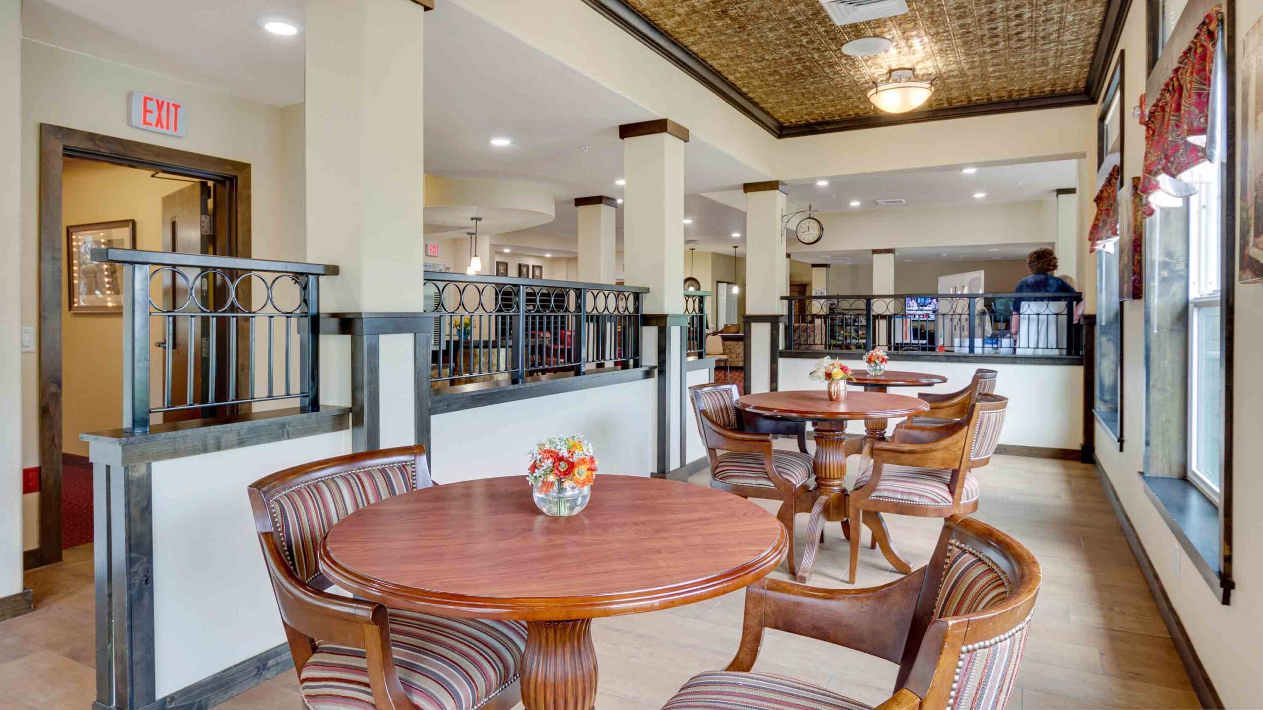 Bright, spacious dining area with round wooden tables and striped chairs. Small flower arrangements sit on each table. Large windows let in natural light, and a few people are visible in the background.