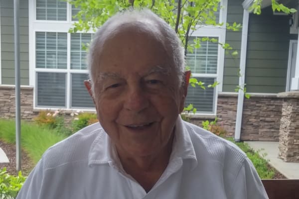 An elderly man with short white hair and a slight smile is sitting outdoors. He is dressed in a white collared shirt. Behind him, there is greenery and a building facade with large windows and stone detailing.