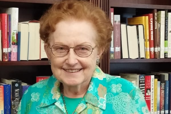 An elderly woman with short, curly red hair and glasses is smiling. She is wearing a turquoise blouse with a floral pattern. Behind her is a bookshelf filled with various books.