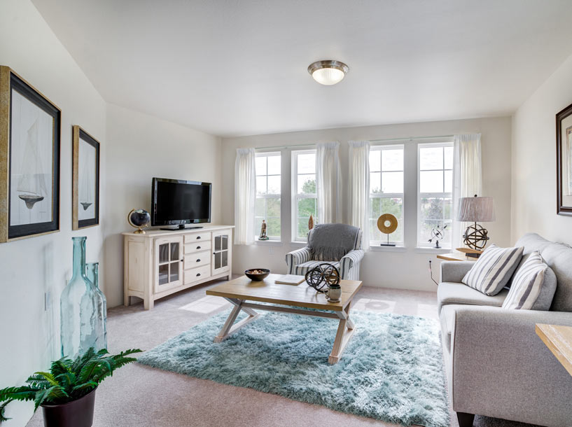 A cozy living room with white walls, featuring a gray sofa, a light wooden coffee table on a blue rug, a TV on a white media console, an armchair, and large windows with sheer curtains. Decorative items including framed art, plants, and nautical accents are displayed.