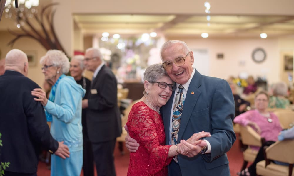 An elderly couple joyfully dances, with the woman in a red dress and the man in a suit and tie, both smiling brightly. Other elderly people socializing and dancing are visible in the background in a warmly lit room.