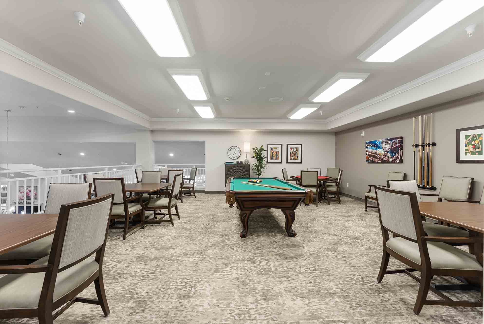 A spacious game room features a pool table with pool cues on the wall, several wooden tables with cushioned chairs around them, framed artwork on the walls, a clock, and overhead fluorescent lighting illuminating the carpeted floor.