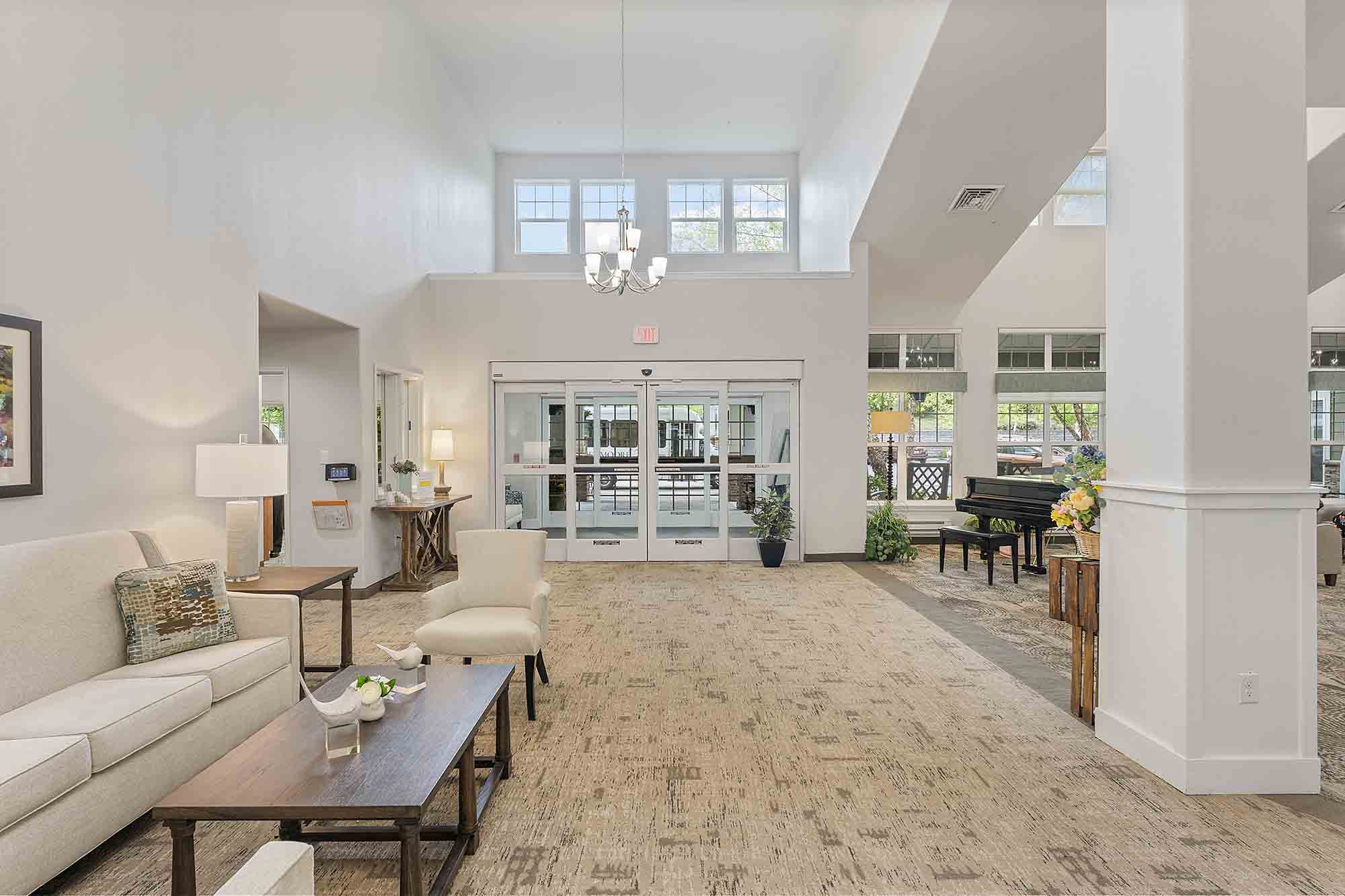 A spacious, well-lit lobby with high ceilings. The area features a white couch, armchairs, and a wooden coffee table. In the background, a grand piano is placed near a window. Double glass doors with sidelights provide access to the outdoors. A chandelier hangs above.