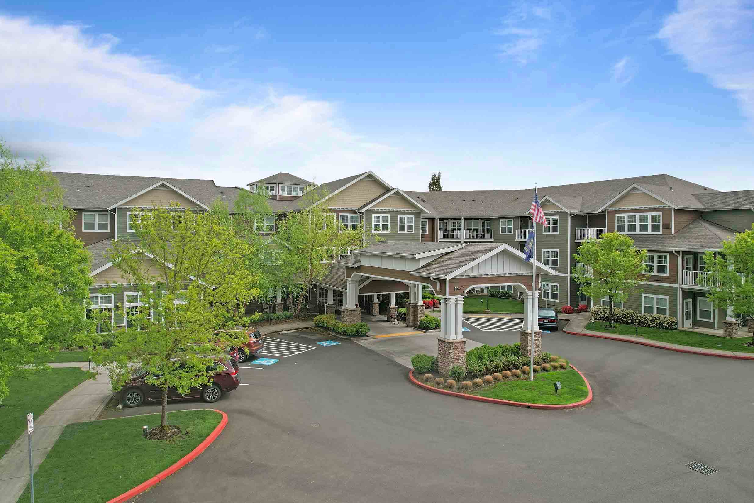 A large, multi-story building with green and beige siding stands under a blue sky. The structure features a covered entrance supported by white pillars, an American flag, and surrounding greenery with well-maintained trees and bushes. Several cars are parked nearby.