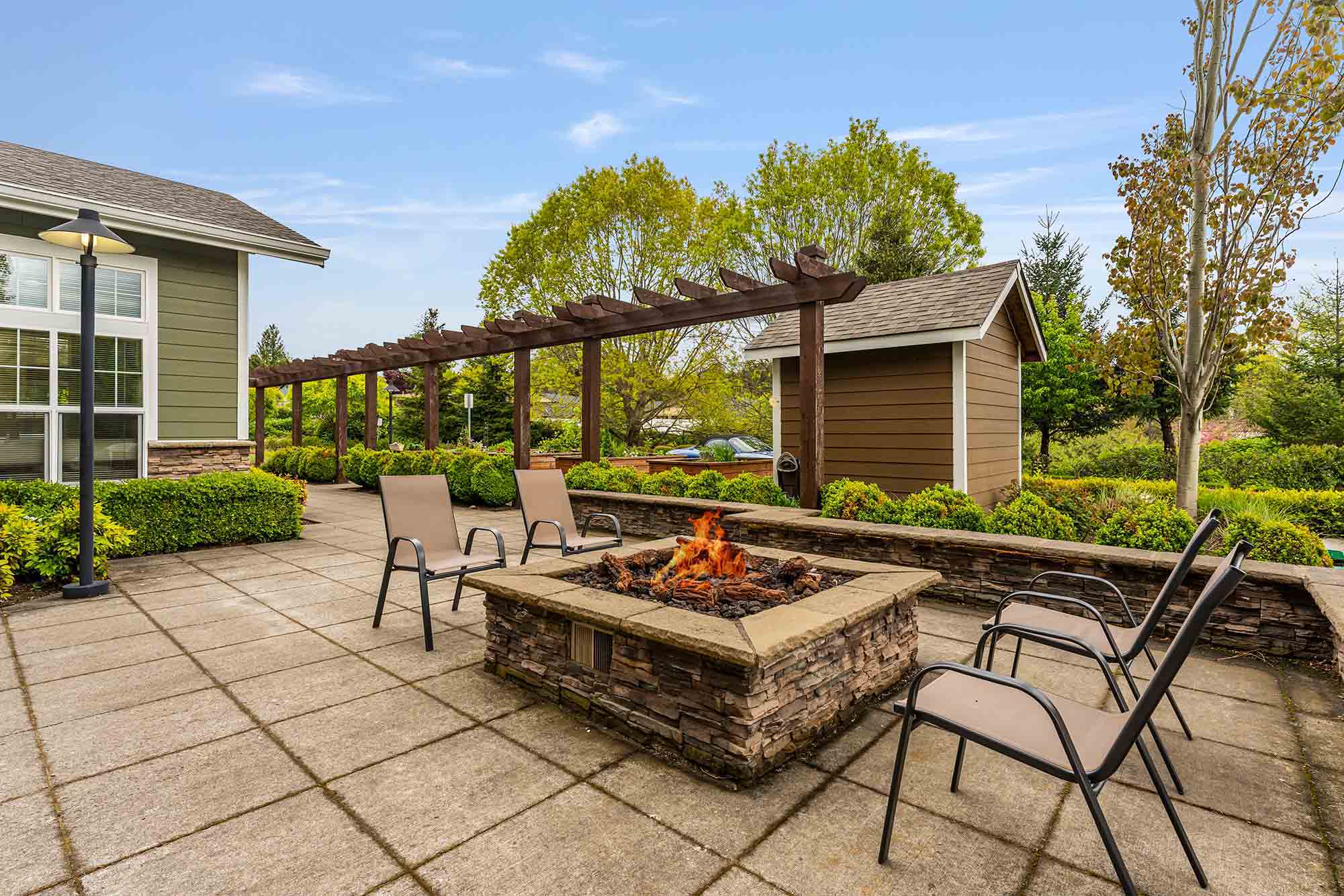 A backyard patio with a fire pit surrounded by four chairs on a tiled surface. A pergola extends from a nearby shed-like structure and a well-maintained garden with hedges and trees in the background. The sky is clear and blue.