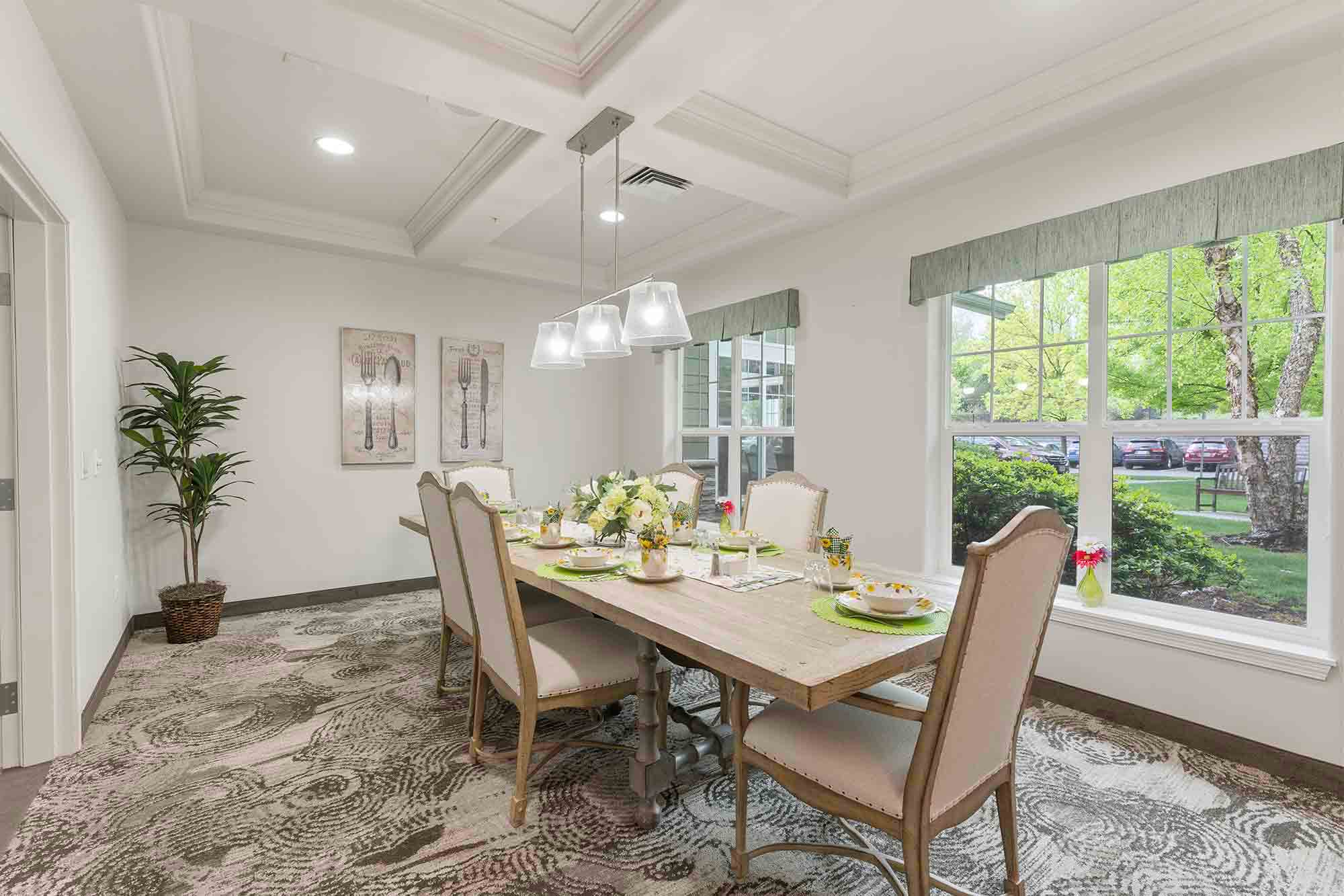 A well-lit dining room with a large wooden table surrounded by six upholstered chairs. The table is set for a meal with plates, glasses, and floral centerpieces. Large windows on one side offer a view of green trees and a parking area. A potted plant and artwork adorn the room.