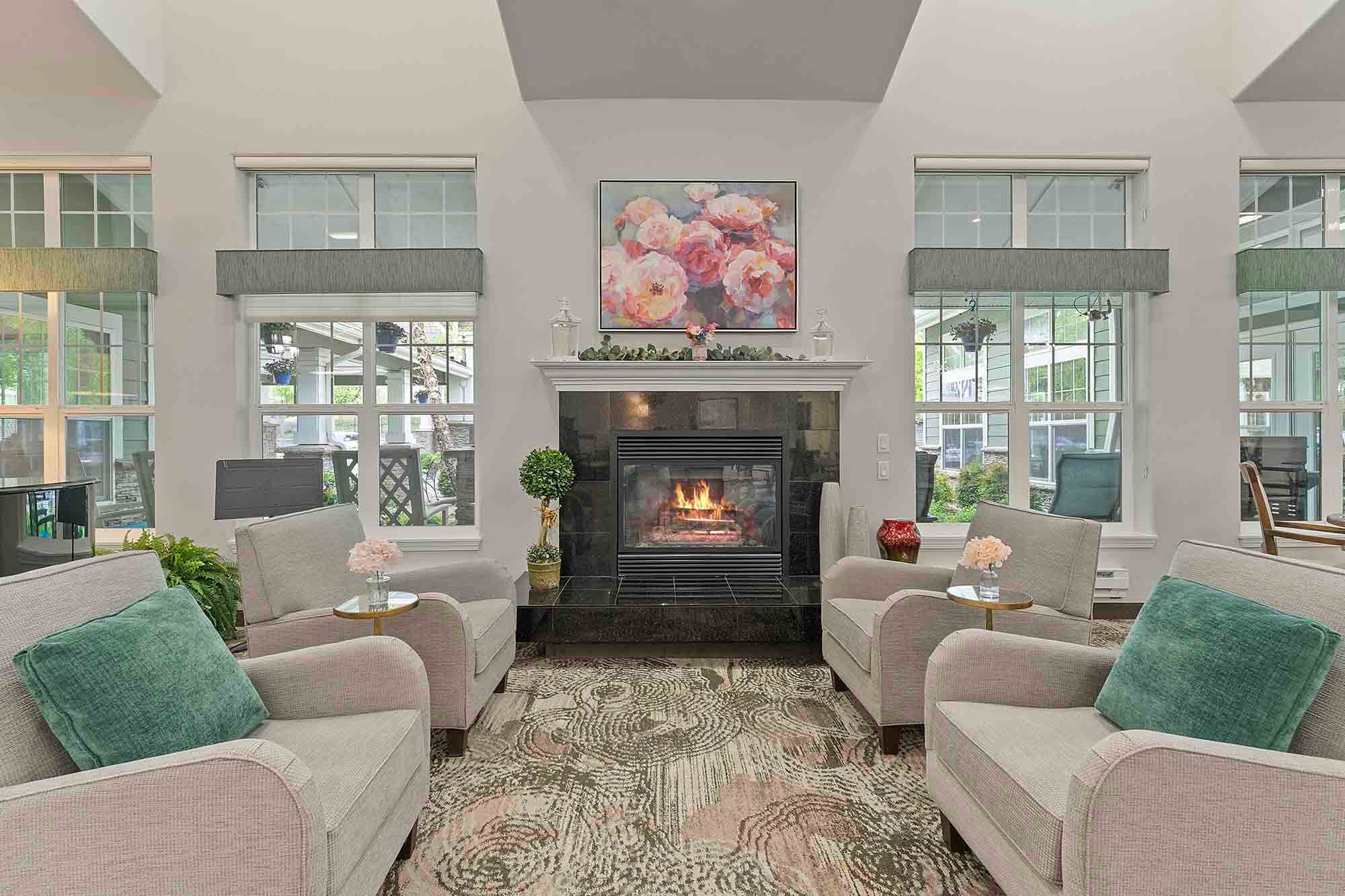 A cozy living room featuring a lit fireplace. Four armchairs with green cushions are placed around a coffee table in front of the fireplace. The walls are adorned with windows and a floral painting above the fireplace. The room has a patterned rug and decor.