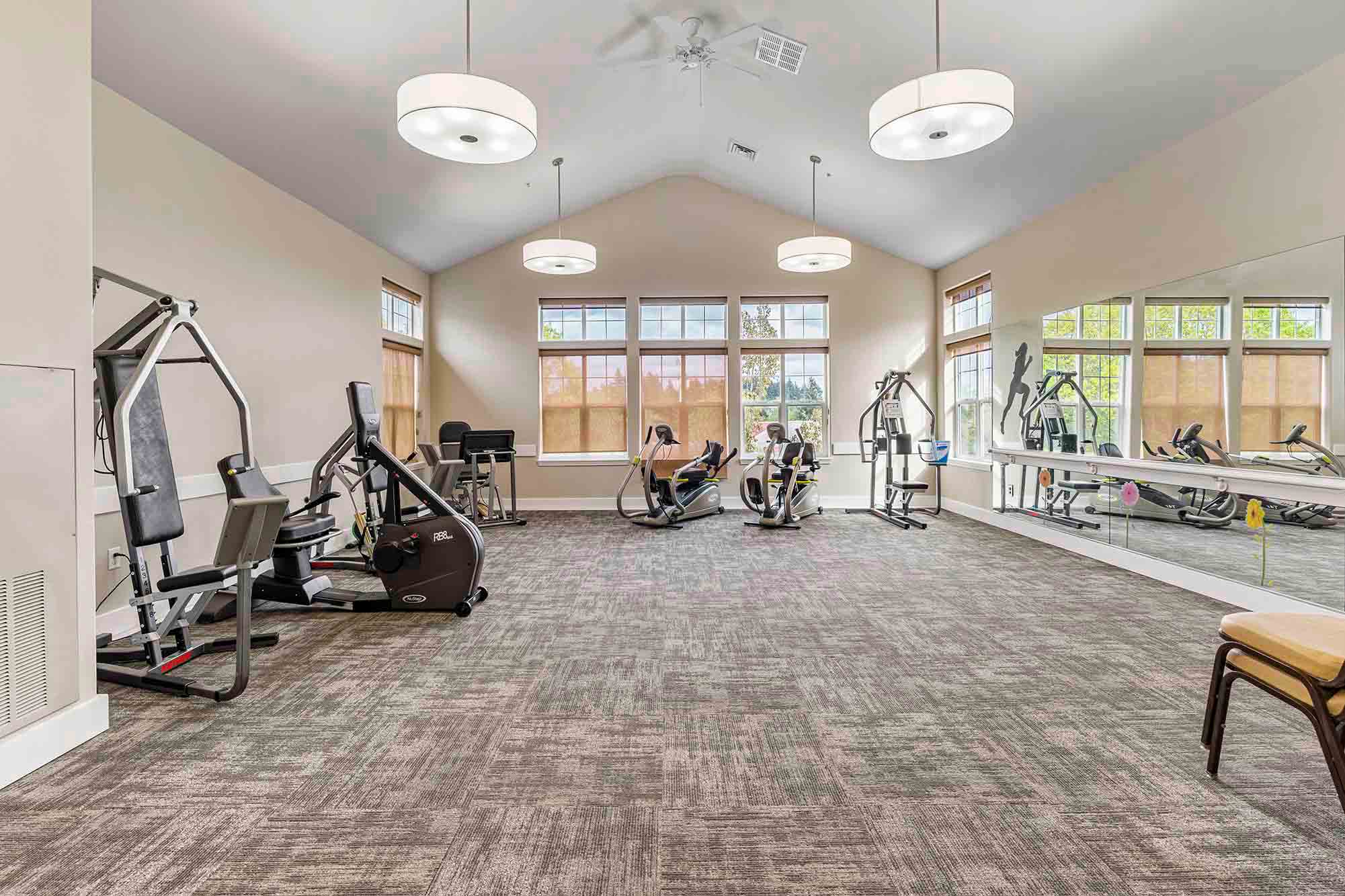 A spacious fitness room with high ceilings, large windows, and various workout machines including stationary bikes, treadmills, and strength training equipment. Mirrors line one wall, and the room features modern lighting with carpeting on the floor.