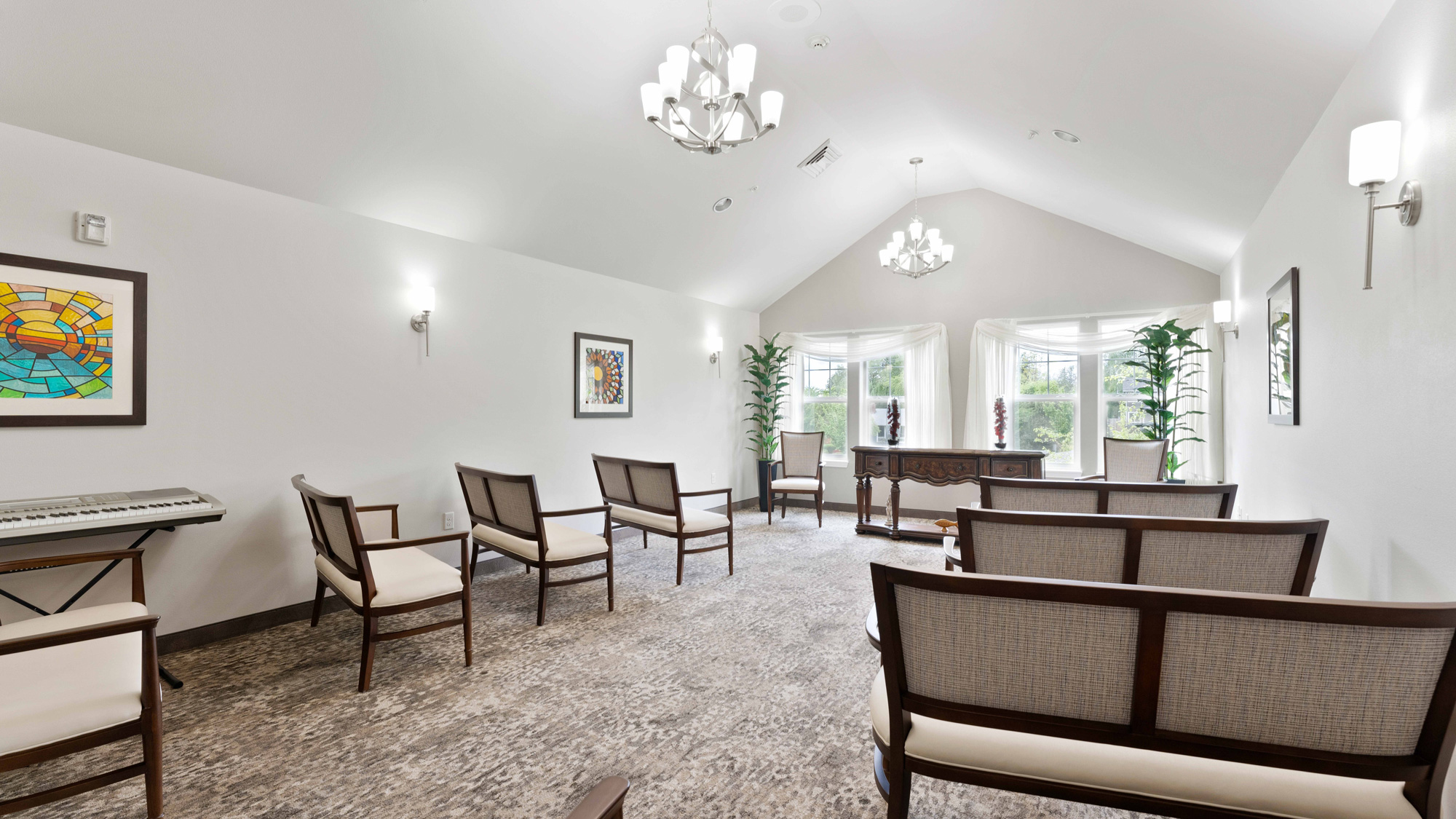 A bright, modern room with white walls, several cushioned chairs, a desk near large windows, wall art, potted plants, a keyboard, and chandeliers hanging from a vaulted ceiling.