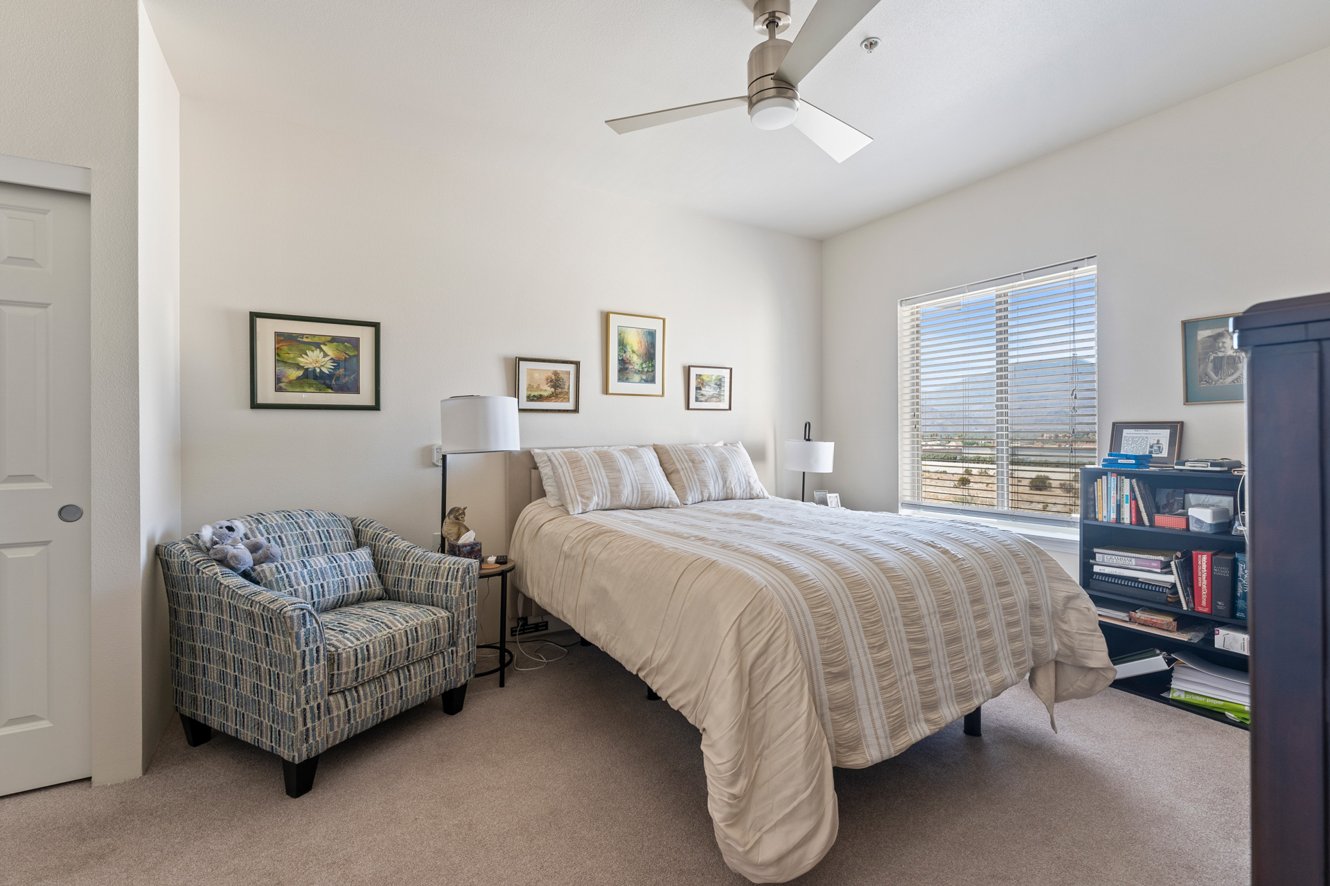 A bright bedroom with a neatly made bed, patterned armchair with a stuffed animal, framed art on white walls, a window with blinds, a ceiling fan, and a bookshelf.