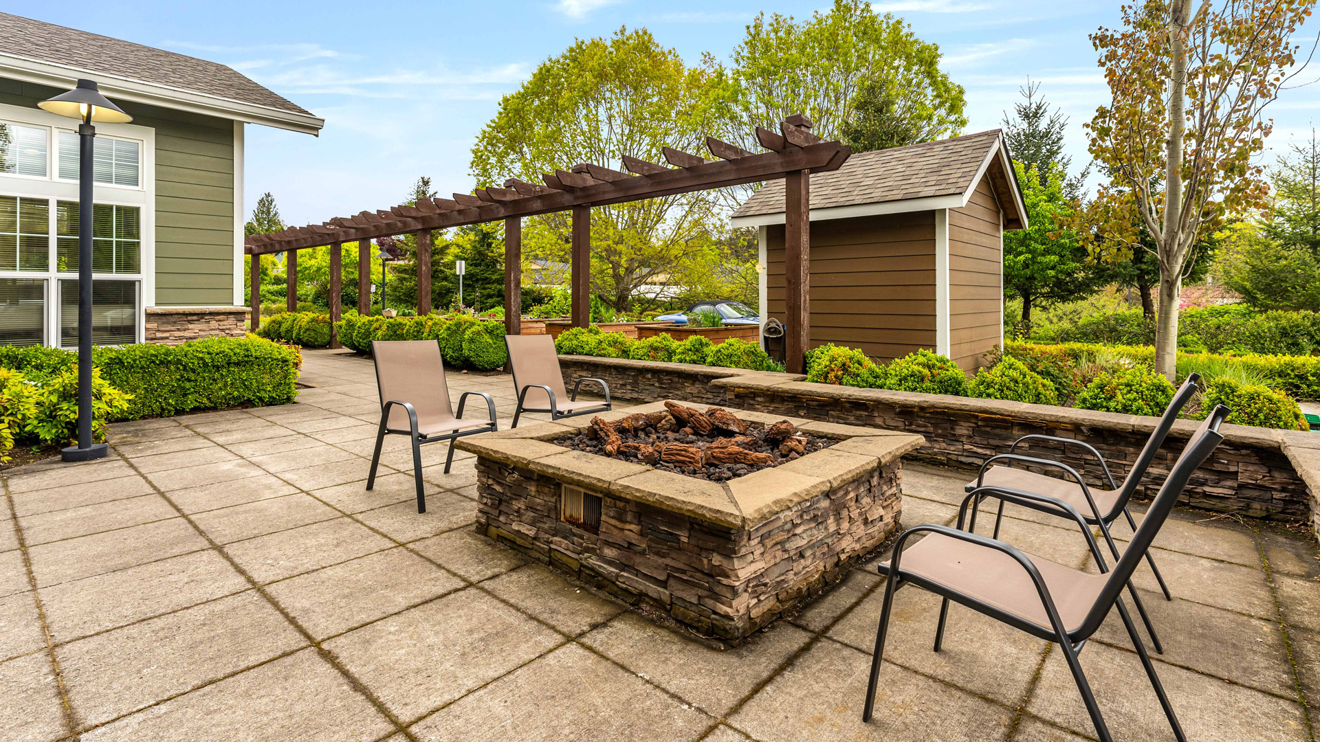 Four patio chairs surround a stone fire pit on a tiled patio, with a wooden pergola, small shed, neatly trimmed hedges, and trees in the background under a blue sky.