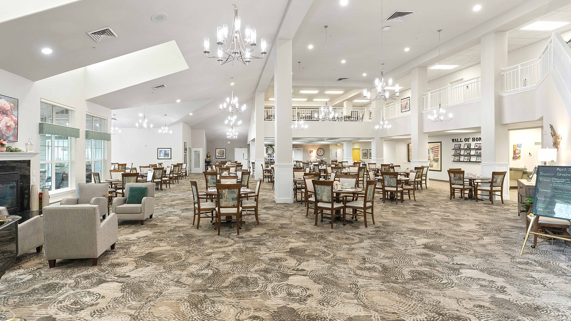 Spacious, well-lit dining area with numerous wooden tables and chairs, chandeliers hanging from the ceiling, and a patterned carpet. There are a few lounge chairs and a fireplace on the left side.