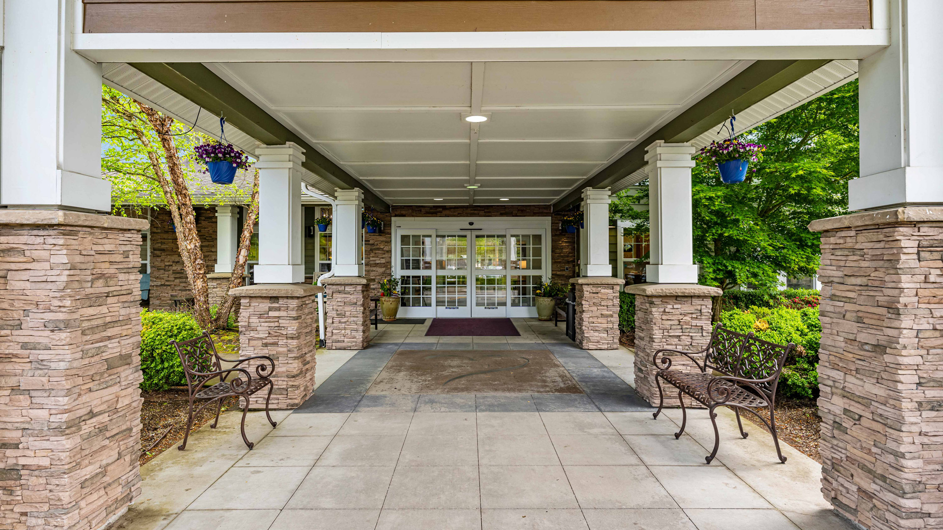 Covered entrance of a building with stone pillars, hanging flower pots, benches on each side, and double glass doors leading inside. Green trees and shrubs surround the walkway.