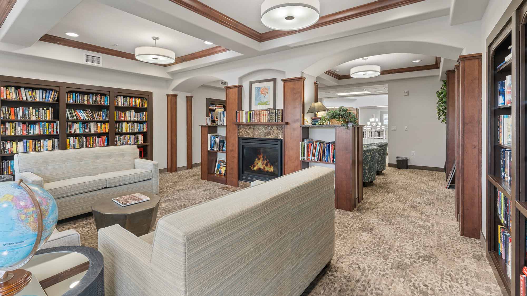 A cozy library room with bookshelves, beige sofas, a fireplace, a globe, and modern lighting. Books line the shelves, and a hallway is visible in the background.