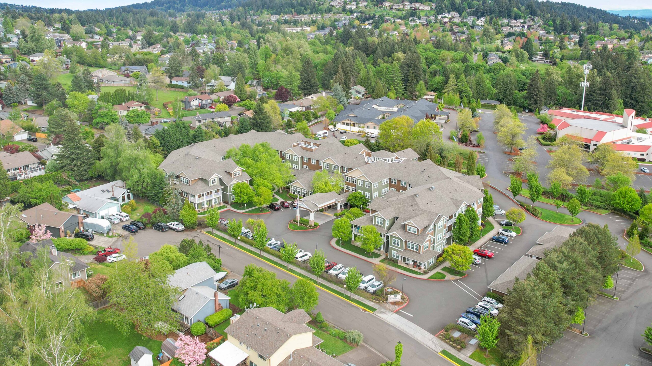 Aerial view of a suburban neighborhood with large buildings, parking lots, houses, and many green trees surrounding the area. The scene appears peaceful and well-maintained.