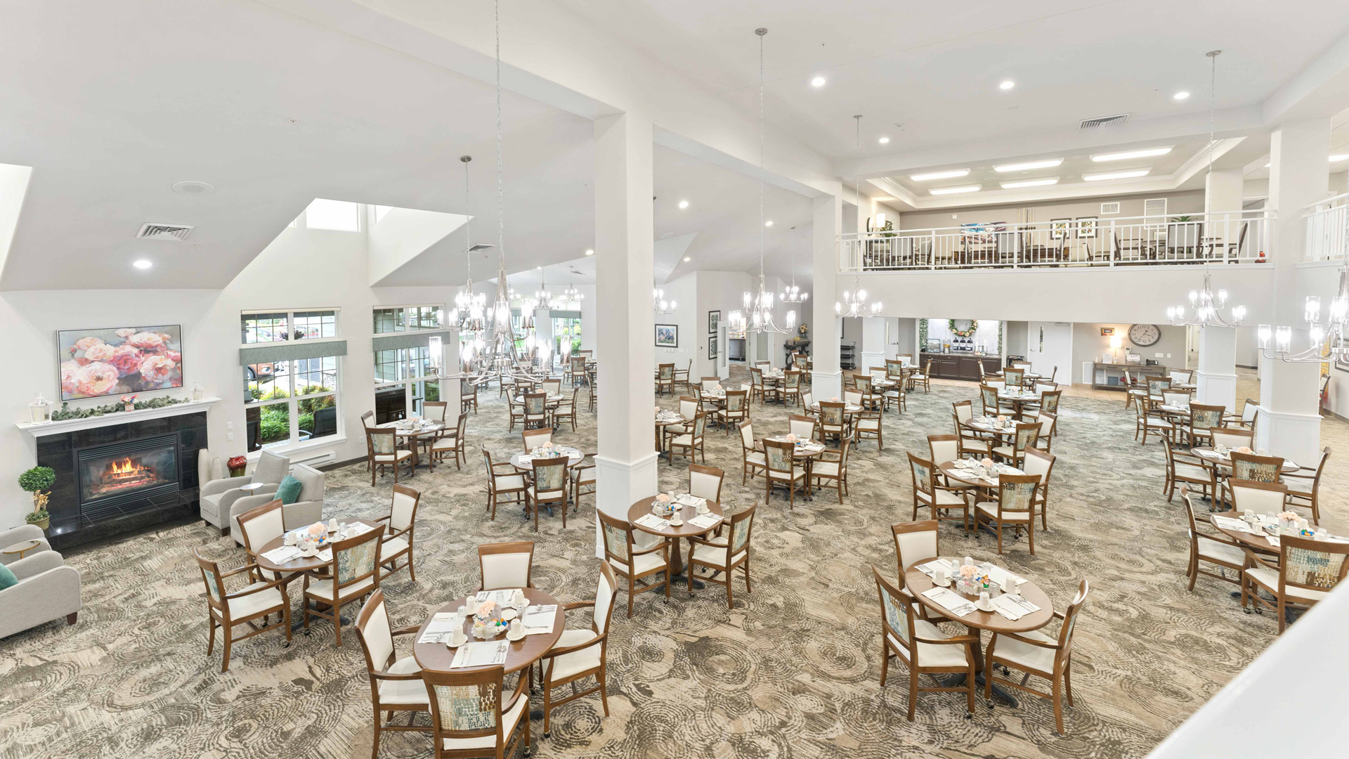 Spacious, elegant dining hall with numerous round tables and chairs, chandeliers, large windows, a fireplace, and a balcony seating area. Neutral tones and tasteful decor create a welcoming atmosphere.