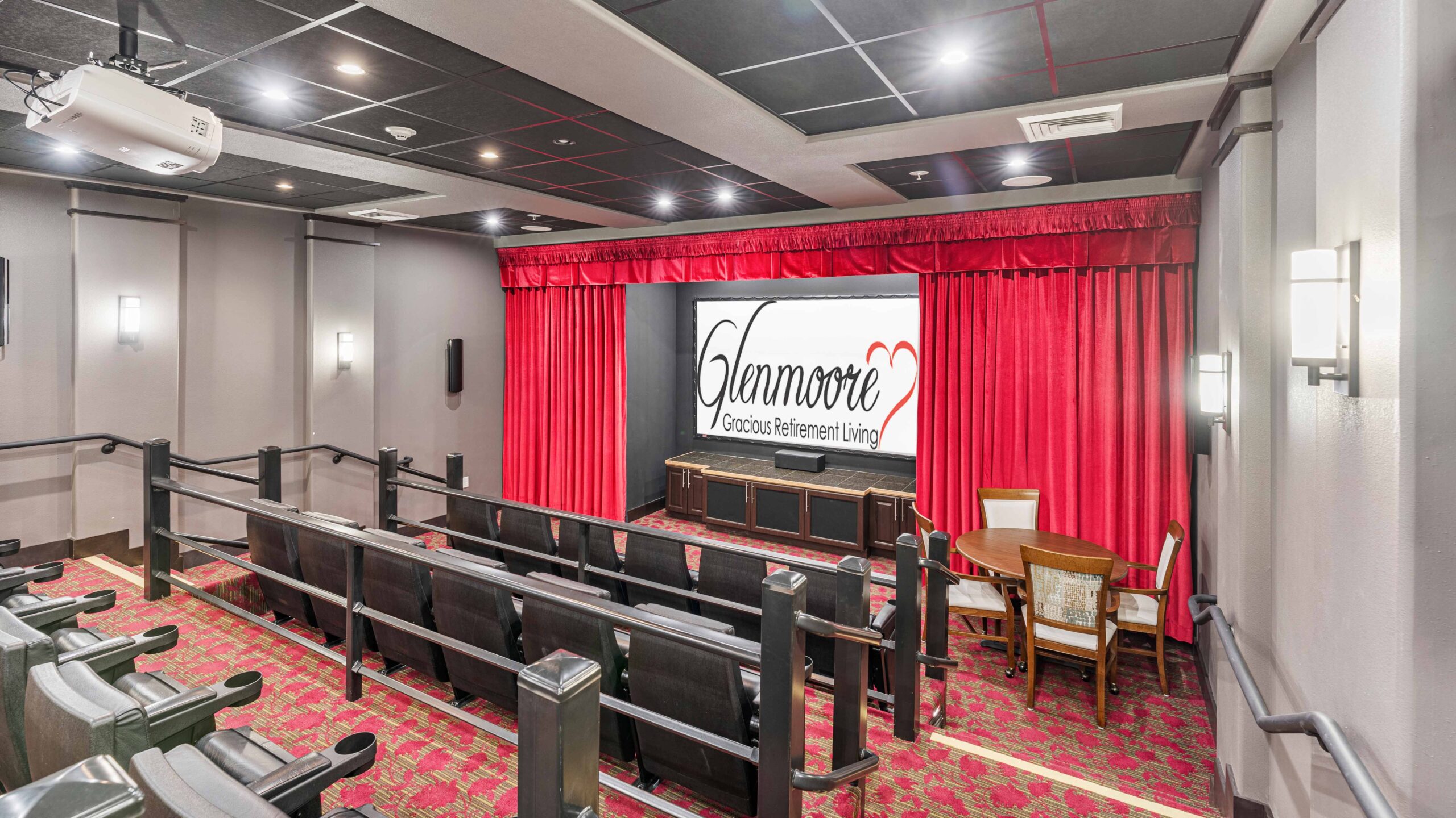 A small movie theater with red carpet and curtains, black chairs, a projector, and a screen displaying "Kenmore Gracious Retirement Living." A table with two chairs sits near the screen. The room has soft wall lighting.