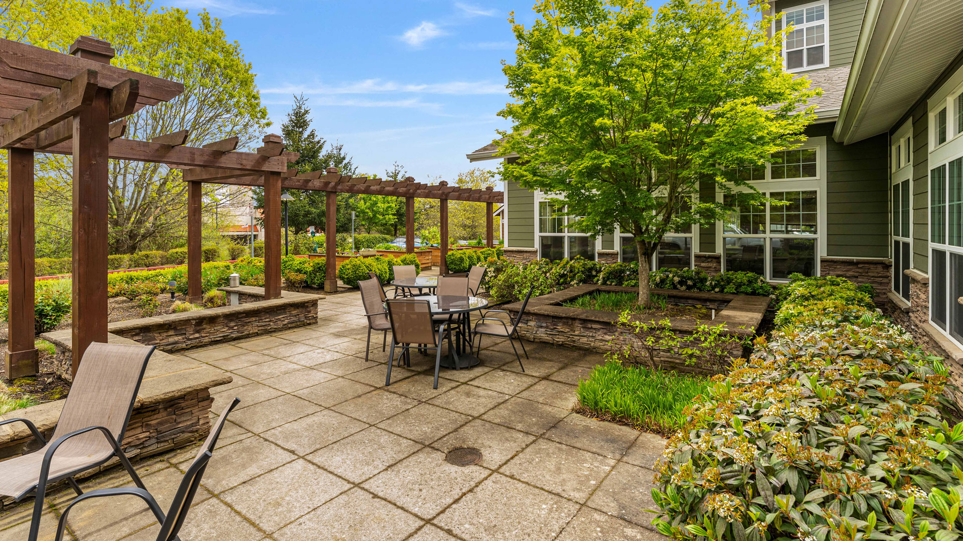 Outdoor patio with metal tables and chairs, surrounded by lush green trees, shrubs, and stone planters. A wooden pergola provides partial shade, and a building with large windows is visible in the background.