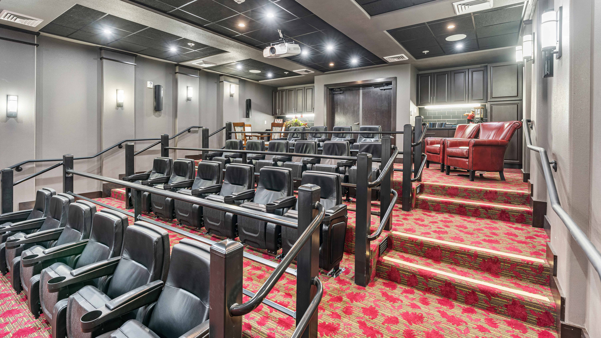 A modern home theater room with tiered seating in black chairs, red patterned carpet, wall-mounted lights, a projector on the ceiling, and a row of two red armchairs at the back near a snack counter and cabinets.