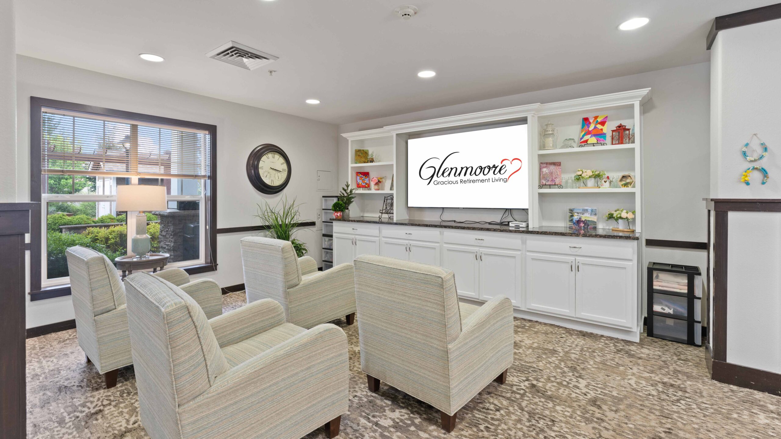 A cozy lounge area with four striped armchairs facing a TV displaying "Glenmore Gracious Retirement Living." Shelves hold books, decor, and flowers; large windows and a clock adorn the walls.