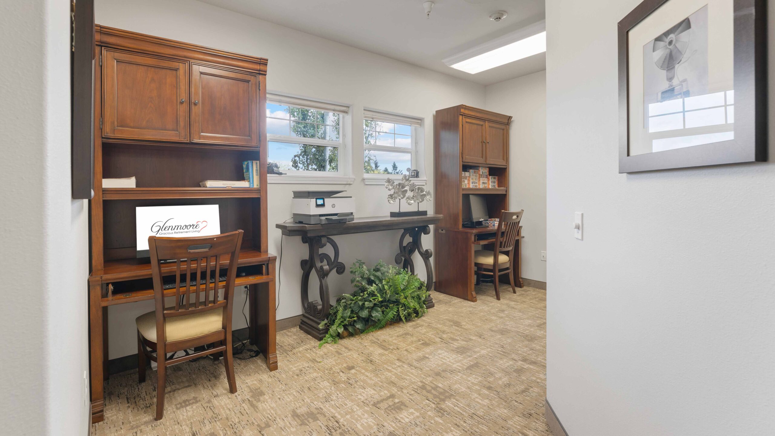 A small office space with wooden desks and chairs, a computer monitor, a decorative plant, shelves with items, and two windows letting in natural light. A framed picture hangs on a white wall in the foreground.
