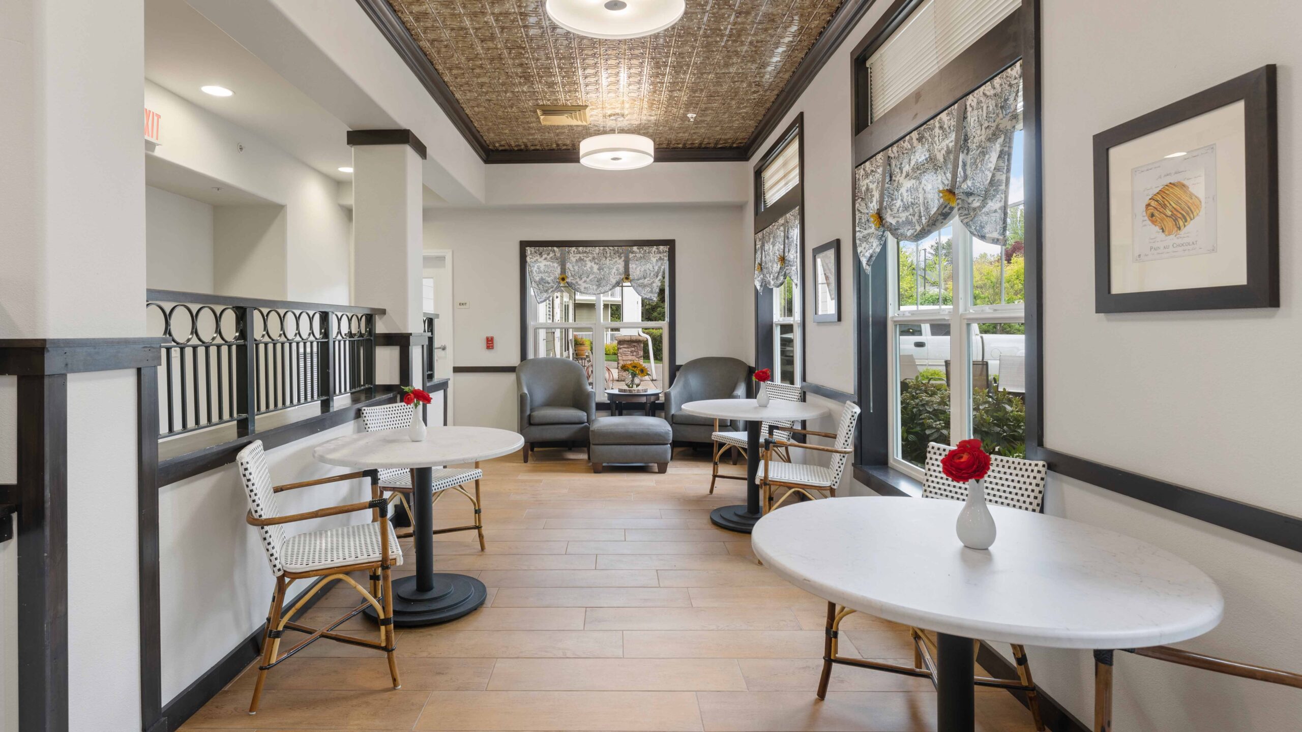 A bright, cozy café area with small round tables, white chairs, gray armchairs by large windows, wood flooring, and simple décor. Each table has a small vase with a red flower.