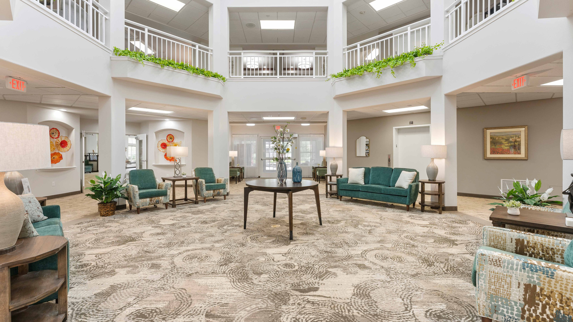 A bright, spacious lobby with modern decor, featuring teal and beige seating, potted plants, round tables with lamps, and a central table with a floral arrangement. White railings and upper balcony overlook the area.
