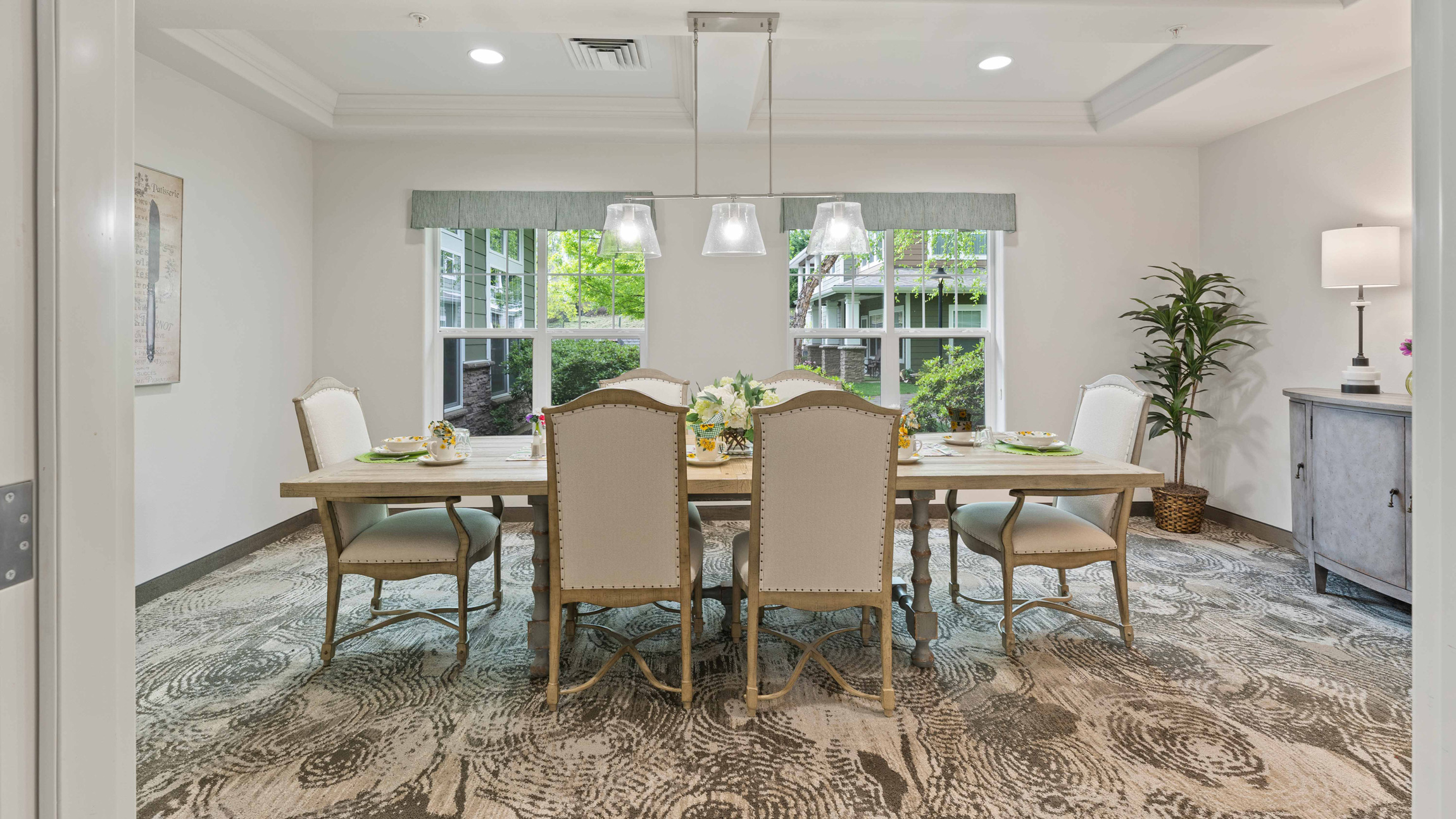 Elegant dining room with a wooden table set for six, upholstered chairs, modern light fixture, large windows showing greenery, patterned carpet, a sideboard, potted plant, and soft, neutral tones throughout.