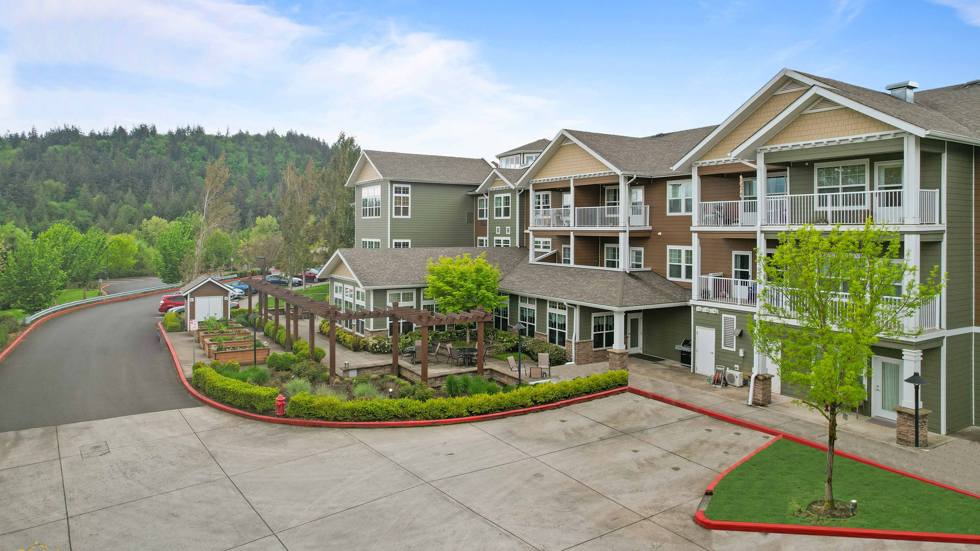 A modern, multi-story apartment building with balconies overlooking landscaped gardens and a pergola-covered walkway, set against a backdrop of green trees and hills under a blue sky.