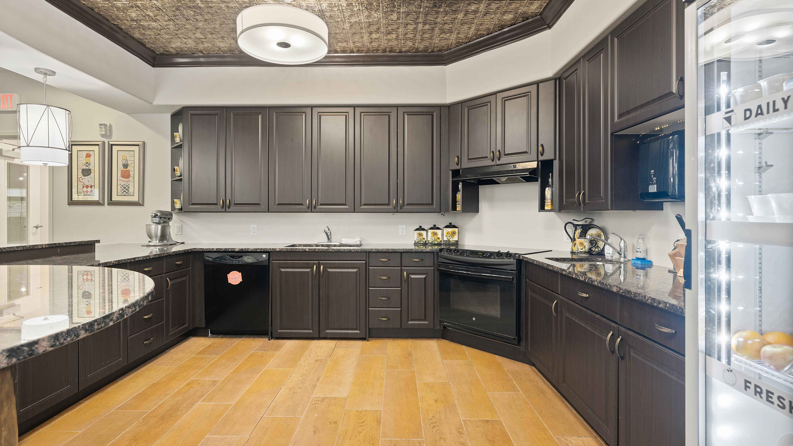 Modern kitchen with dark wood cabinets, granite countertops, stainless steel appliances, a black oven, and a wood-look tile floor. A fridge with glass doors and labeled shelves is partially visible on the right.