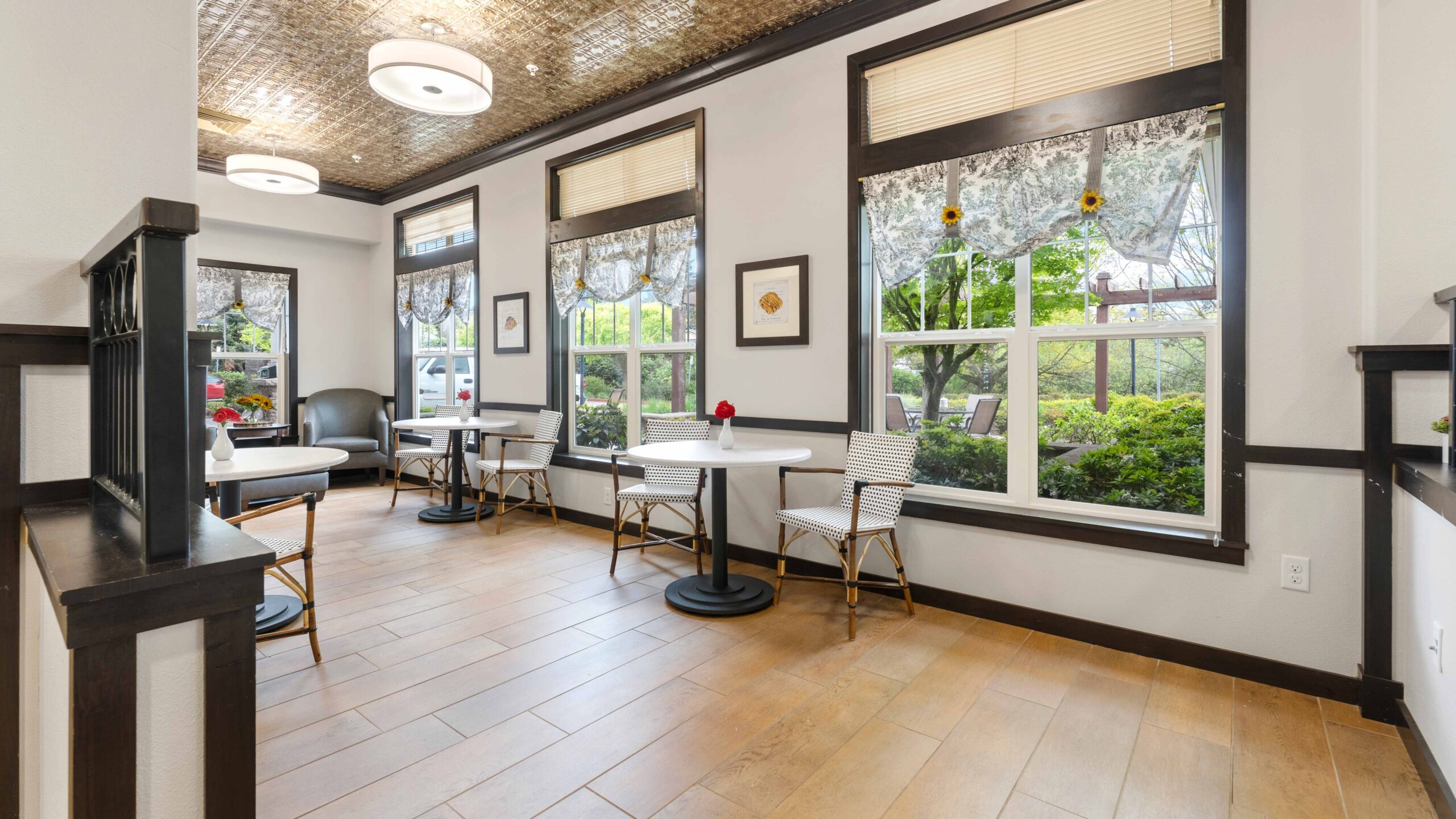 A bright café area with large windows, white tables, and chairs. Floral lace curtains and framed art decorate the walls. Greenery is visible through the windows, and the space has wooden floors and a patterned ceiling.