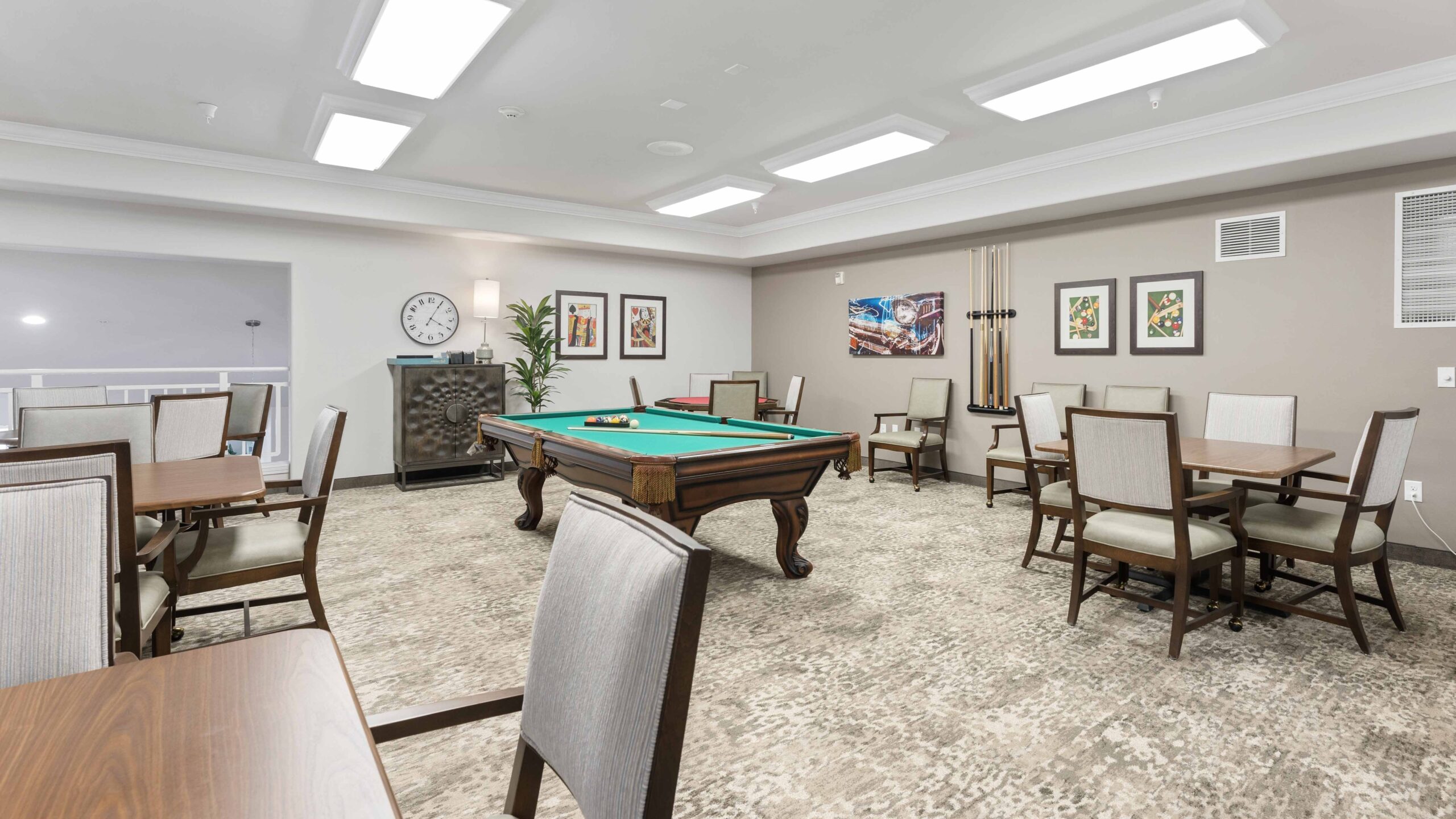 Spacious game room with a pool table in the center, surrounded by tables and chairs. Art decorates the walls, and cues are mounted nearby. Bright ceiling lights illuminate the neutral-toned room.