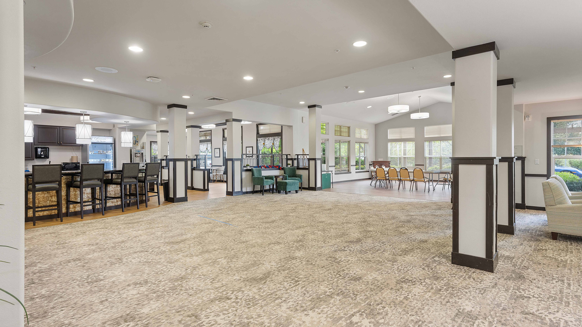 Spacious, well-lit community room with carpeted floors, dining tables, chairs, modern light fixtures, and large windows letting in natural light. A kitchen area with barstools is visible in the background.