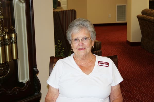 An elderly woman with short gray hair and glasses sits smiling in a room with red carpet. She is wearing a white shirt and a red name tag. A tall wooden structure with gold details is visible to her left.
