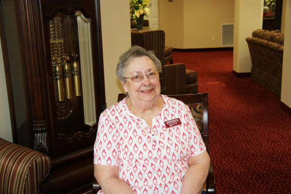 An elderly woman with glasses and short hair, wearing a white and red patterned shirt and a name badge, is sitting in a chair with a warm smile. The background features a carpeted hallway and a grandfather clock.