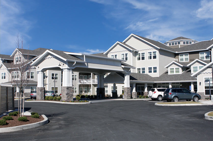 Large multi-story building with light-colored exterior and peaked roofs, resembling a modern residential or assisted living facility. Entrance has a covered drop-off area supported by stone columns. Surrounding landscaping includes small shrubs and trees.
