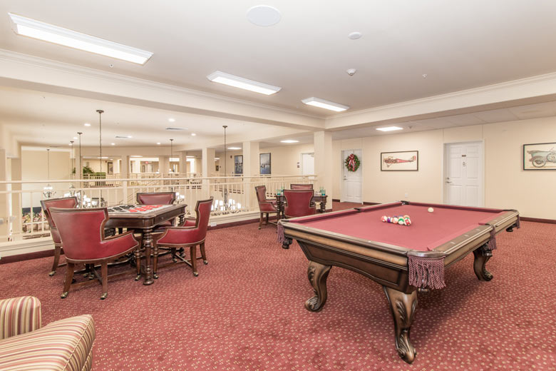 A well-lit recreation room with red carpet features a wooden pool table with red felt and arranged balls, two round tables with red-cushioned chairs for card games, and framed artwork on the walls. Chandeliers hang from the ceiling, illuminating the space.