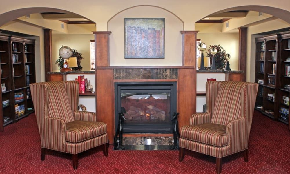 A cozy library with two striped armchairs facing a fireplace, flanked by wooden bookshelves filled with books and decorative items. The red carpet and arched doorway add to the warm ambiance, and a colorful abstract painting hangs above the fireplace.