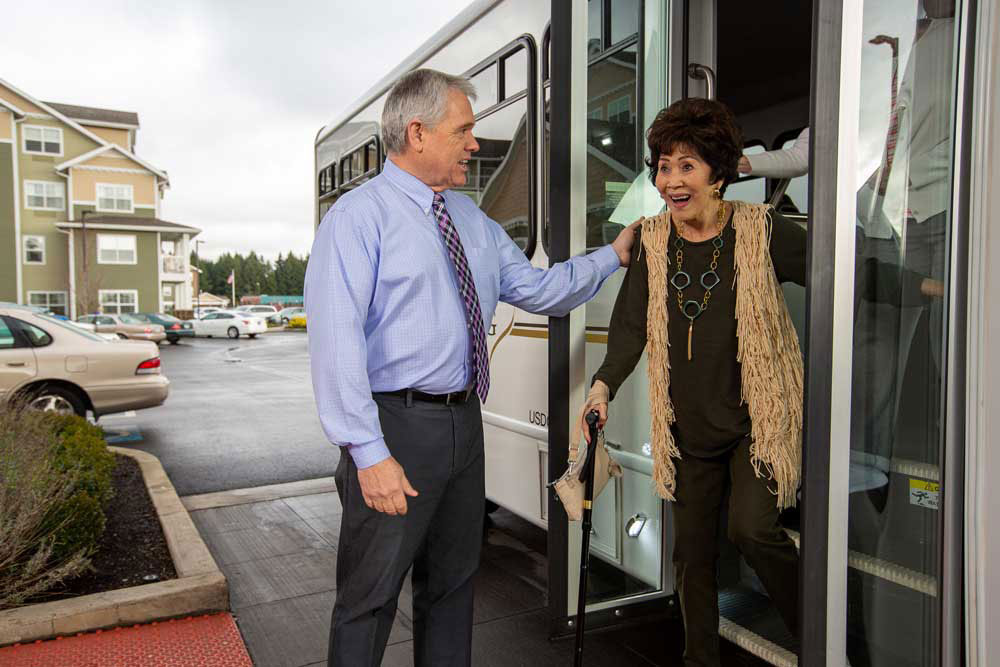 A smiling senior woman, holding a cane and wearing a fringed vest, exits a minibus. A gentleman in a blue shirt and tie stands beside her, offering assistance with a supportive hand on her shoulder. In the background, parked cars and a building are visible.