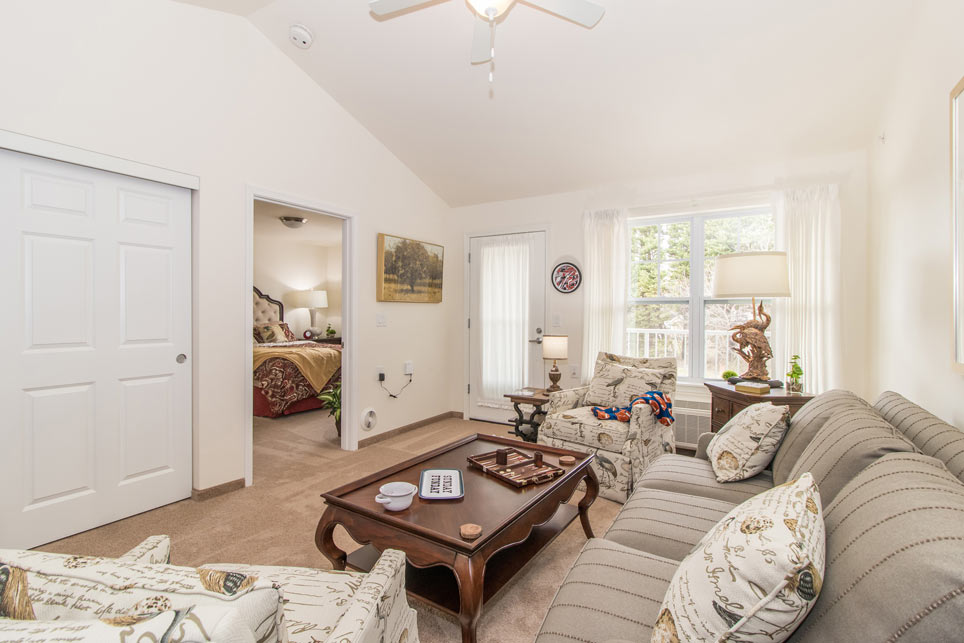 A cozy living room with neutral tones featuring a gray sofa, two patterned armchairs, and a wooden coffee table with decorative items. A bedroom is visible through an open door. The room is illuminated by natural light from a large window and a glass door.