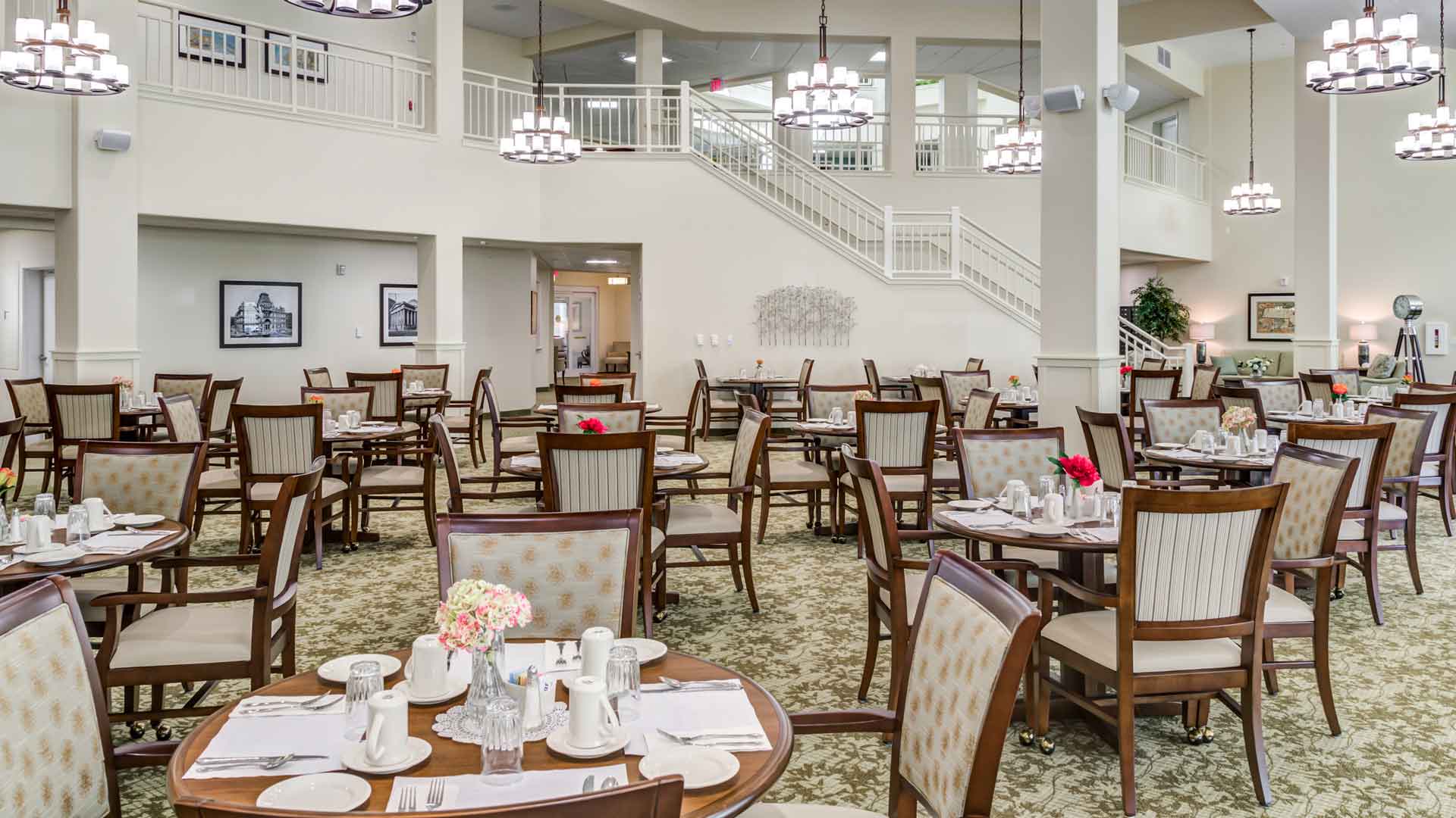 A spacious dining area with neatly arranged wooden tables and chairs, set with white tableware and red flower centerpieces. Chandeliers hang from the high ceiling, and a staircase is visible in the background. Walls feature framed pictures.