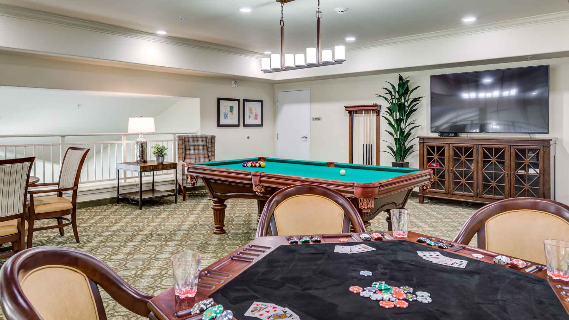 A recreation room with a pool table, billiard balls set up, a poker table with cards and chips, four chairs, and a large flat-screen TV on a cabinet. There are framed pictures, a plant, and a lamp in the background.