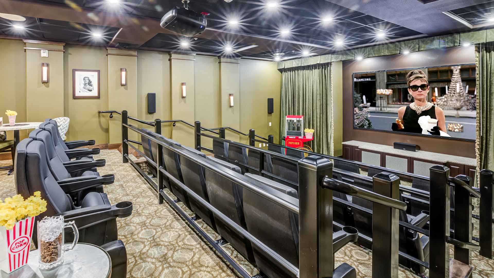 A small movie theater with plush black reclining seats arranged in rows, facing a large screen showing a classic film scene. Popcorn is on a side table, and the room has wall sconces and framed art on the walls.