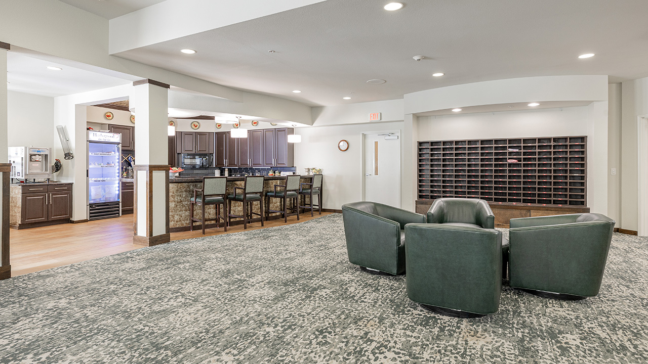 Spacious lobby area with four green armchairs arranged in a circle, a bar with barstools and kitchen area in the background, and a wall of mail slots to the right. The room has neutral tones and modern decor.