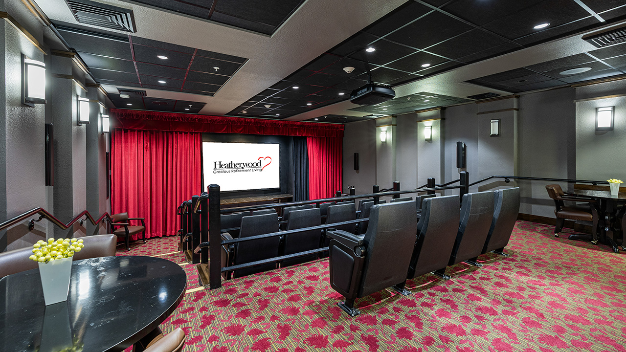 A small theater room with rows of leather seats facing a screen, red curtains, decorative wall sconces, and round tables with chairs. The screen displays the logo for Hearthwood Senior Living.