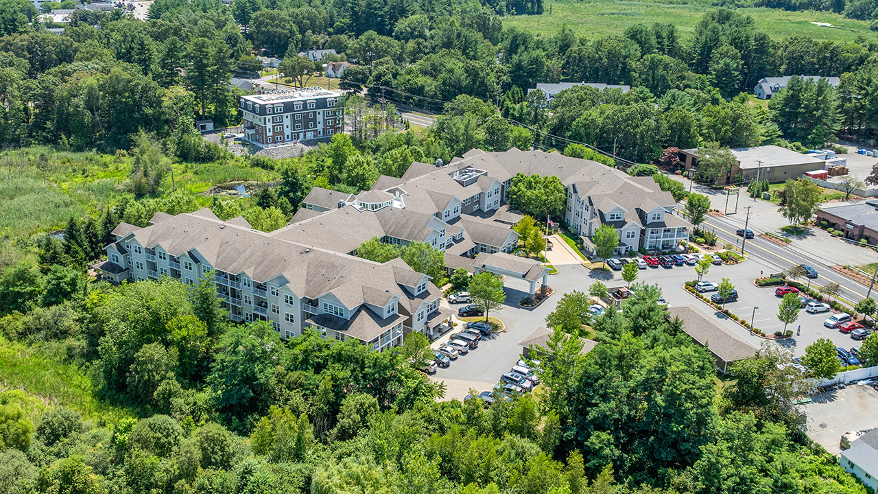Aerial view of a large residential complex surrounded by trees and greenery, with several parked cars and nearby roads, and additional buildings visible in the background.