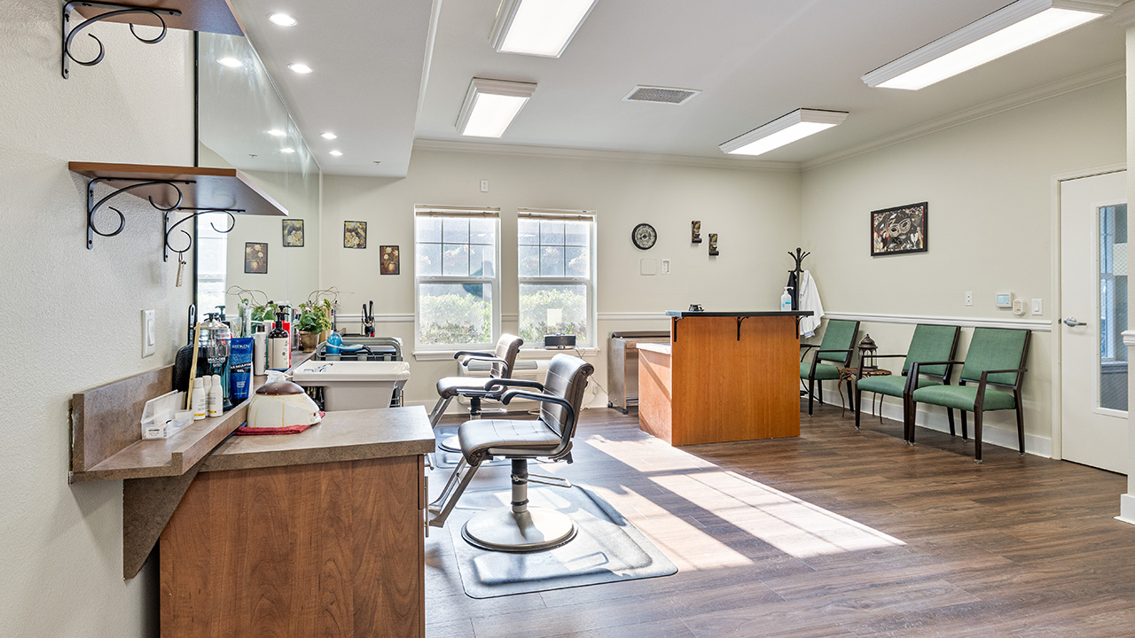 A bright, clean hair salon with two barber chairs, a counter with hair products, a waiting area with green chairs, a wooden reception desk, and large windows letting in natural light.