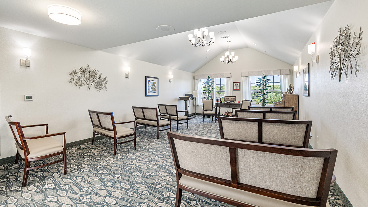 A bright, spacious room with upholstered chairs arranged in rows facing a podium. The room features patterned carpet, wall art, large windows with views of greenery, and modern light fixtures on the ceiling and walls.