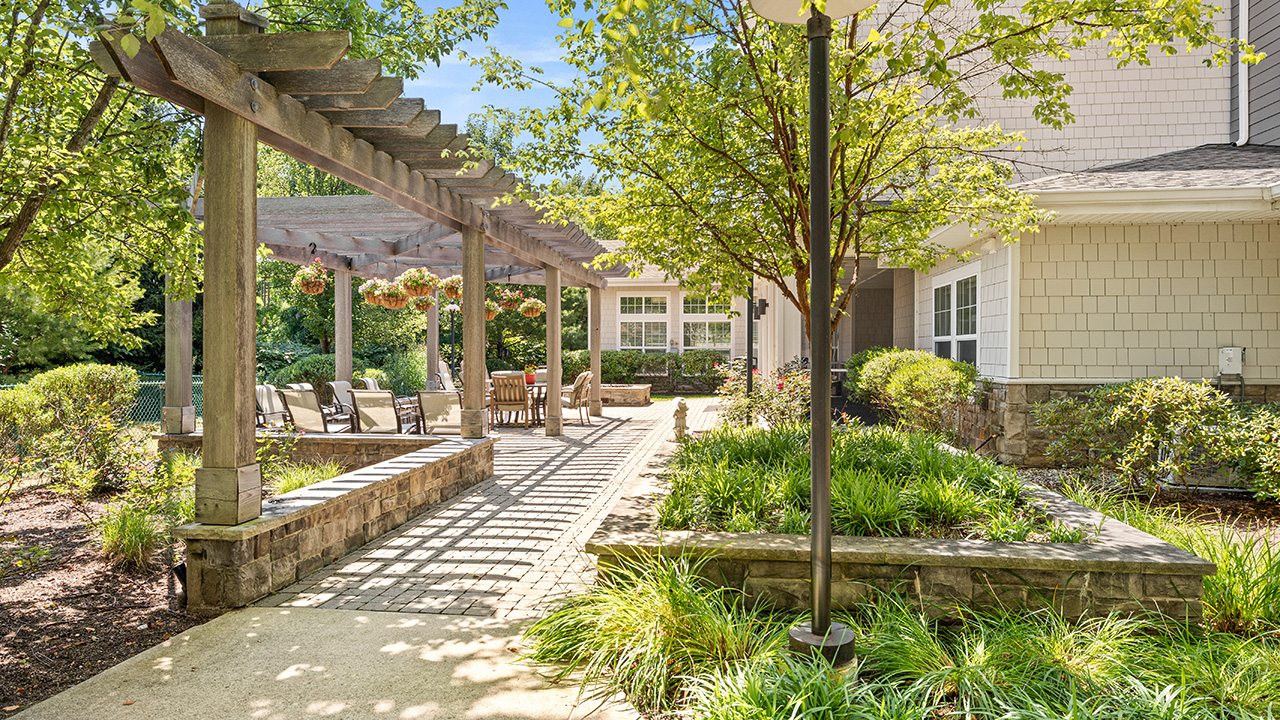 A sunlit outdoor patio with a wooden pergola, hanging flower baskets, dining tables, and chairs, surrounded by green plants and trees next to a beige house.