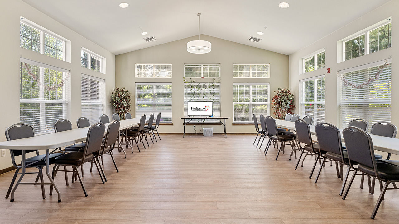 A bright, spacious meeting room with large windows, two rows of tables and chairs facing a screen that displays the word "Welcome." The room has wood flooring and decorative plants by the windows.